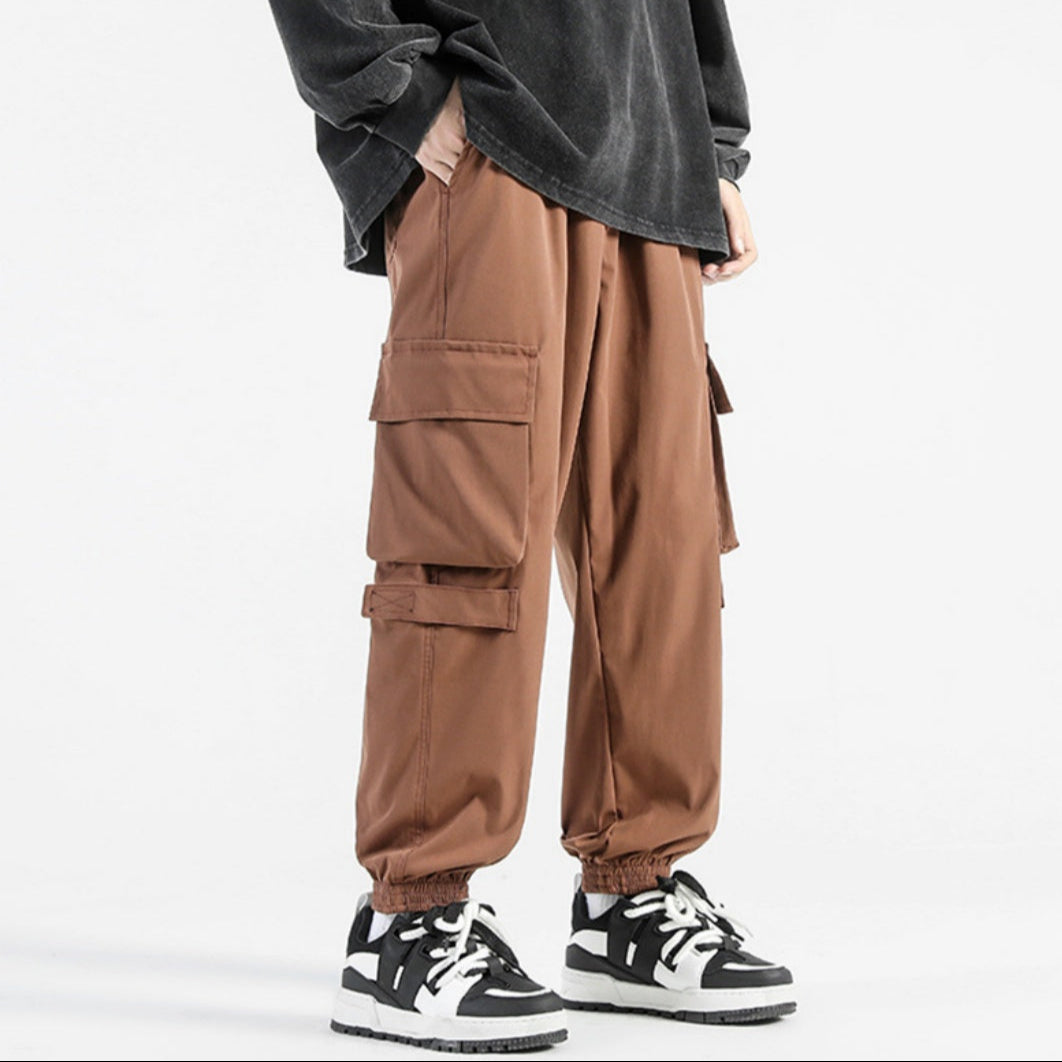 Pantaloni cargo marroni da uomo con tasche multiple, abbinati a sneakers bianche e nere, su sfondo bianco. Moda casual e streetwear.