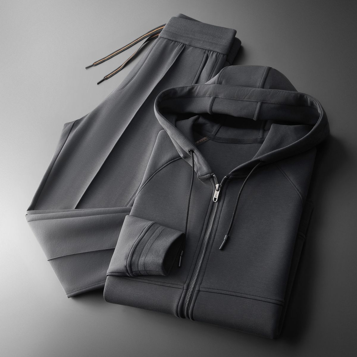 Tuta sportiva grigia in tessuto morbido, composta da felpa con cappuccio e pantaloni con coulisse, su sfondo grigio. Abbigliamento casual elegante.