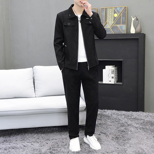 Uomo in tuta sportiva nera, giacca e pantaloni, in piedi su tappeto grigio, accanto a divano bianco e libreria moderna. Moda casual maschile.