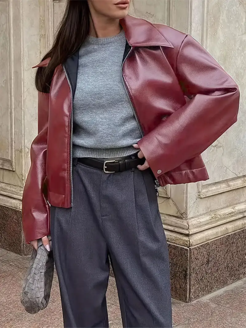 Donna con giacca di pelle rossa, maglione grigio e pantaloni blu scuro, tiene una borsa intrecciata grigia. Moda autunnale elegante e moderna.