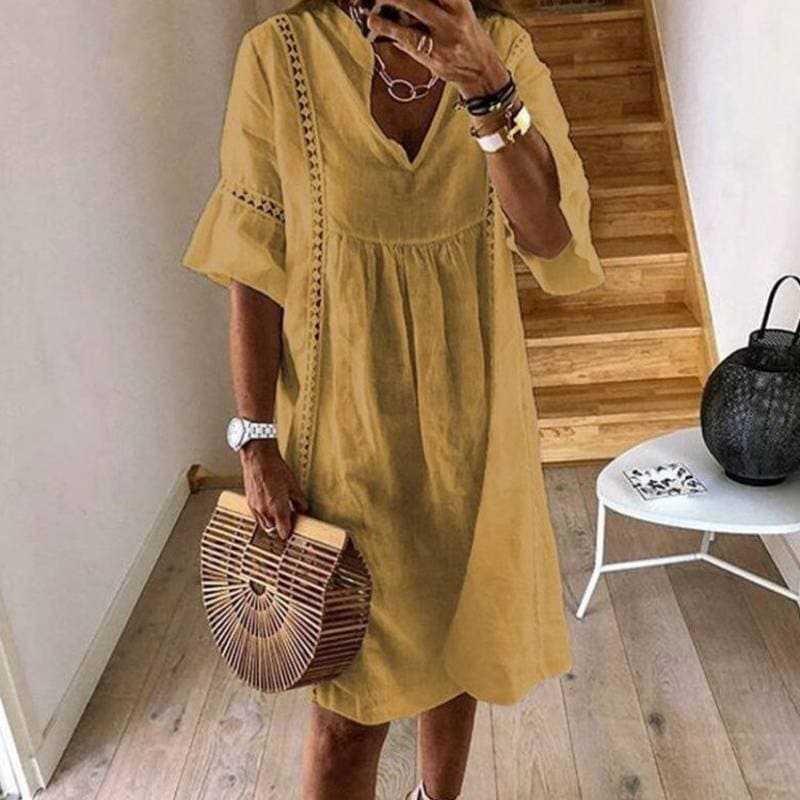 Donna con abito estivo giallo, borsa in legno a mezzaluna, orologio bianco, collane, in piedi su pavimento in legno vicino a scala.