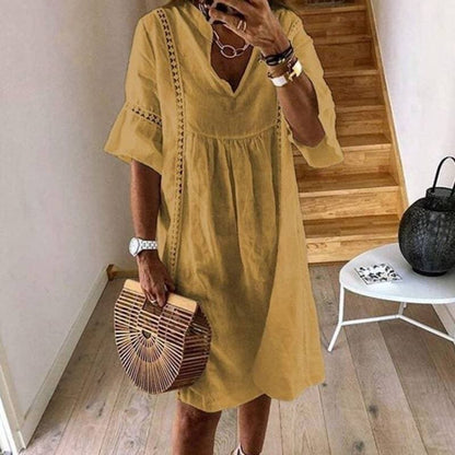 Donna con abito estivo giallo, borsa in legno a mezzaluna, orologio bianco, collane, in piedi su pavimento in legno vicino a scala.