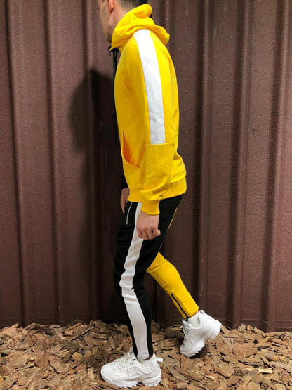 Uomo in tuta sportiva gialla e nera con strisce bianche, scarpe da ginnastica bianche, su sfondo marrone. Moda casual, abbigliamento sportivo uomo.