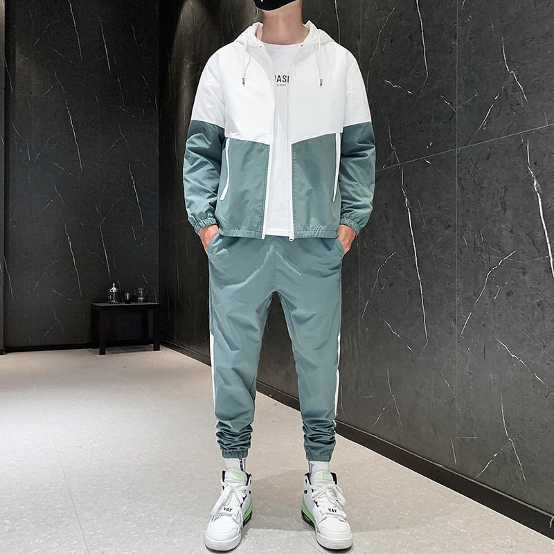 Tuta sportiva uomo verde e bianca, giacca con cappuccio e pantaloni abbinati, scarpe da ginnastica bianche, sfondo parete nera moderna.