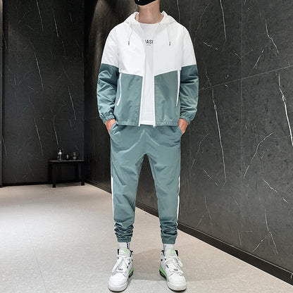 Tuta sportiva uomo verde e bianca, giacca con cappuccio e pantaloni abbinati, scarpe da ginnastica bianche, sfondo parete nera moderna.