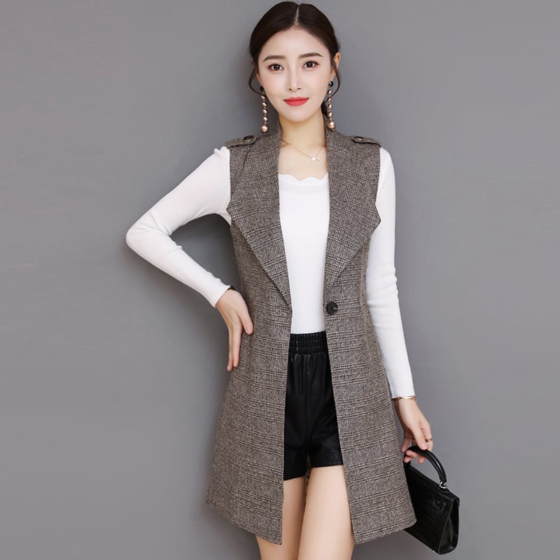 Donna con gilet lungo grigio, maglia bianca e pantaloncini neri, posa su sfondo grigio. Moda autunnale elegante, abbigliamento casual chic.