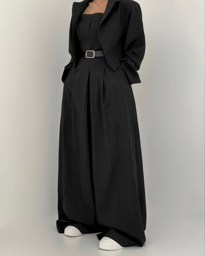 Abito nero elegante a righe verticali con giacca abbinata e cintura, indossato con scarpe bianche. Moda donna, stile sofisticato e moderno.