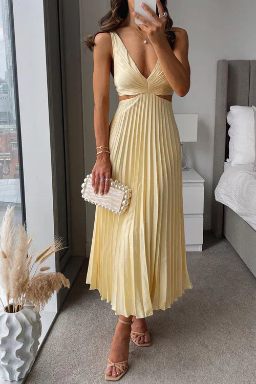 Donna in abito lungo giallo plissettato con scollo a V, sandali eleganti e borsa bianca con perle, in una camera moderna con vista sulla città.