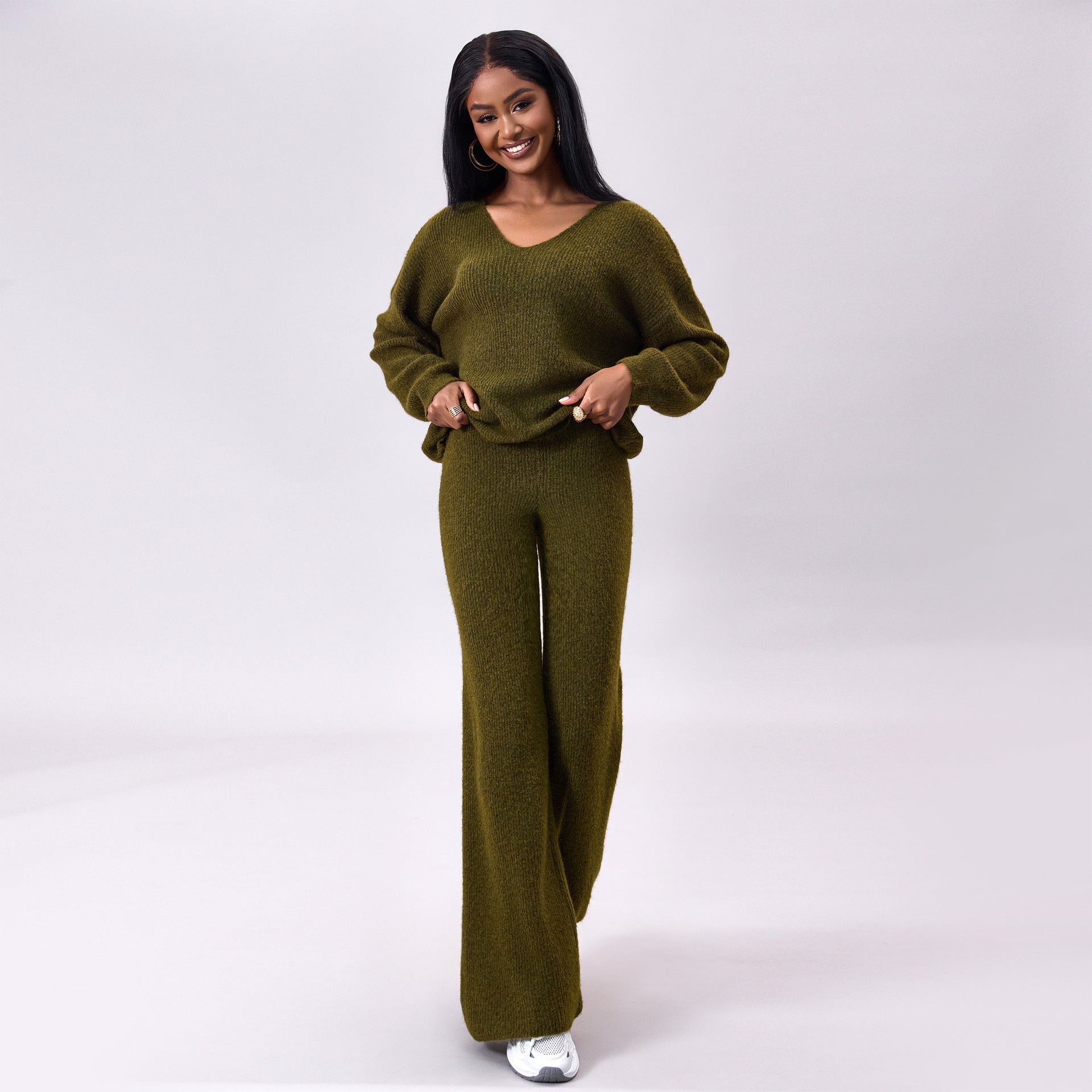 Donna sorridente in abbigliamento coordinato verde oliva, maglione e pantaloni larghi, su sfondo bianco. Moda autunnale, stile casual elegante.