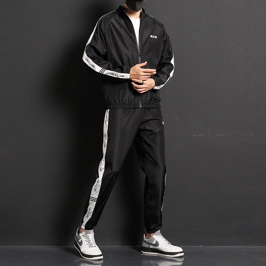 Tuta sportiva nera da uomo con strisce bianche, giacca con zip e pantaloni abbinati, scarpe da ginnastica grigie e bianche, sfondo nero.