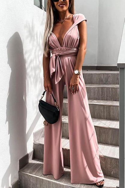 Donna in tuta elegante rosa con scollo a V profondo, cintura annodata, borsa nera e orologio, su scala moderna. Moda femminile chic.