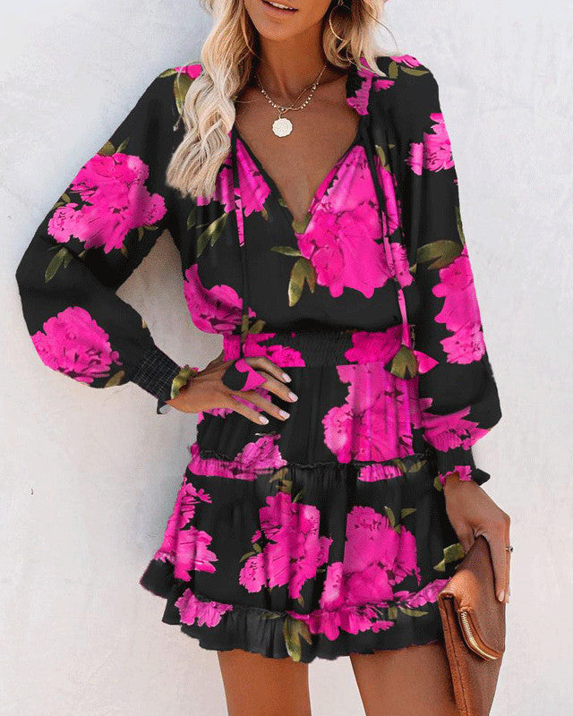 Donna con abito floreale nero e fucsia, maniche lunghe, scollo a V, gonna a balze, borsa marrone. Moda femminile elegante e colorata.