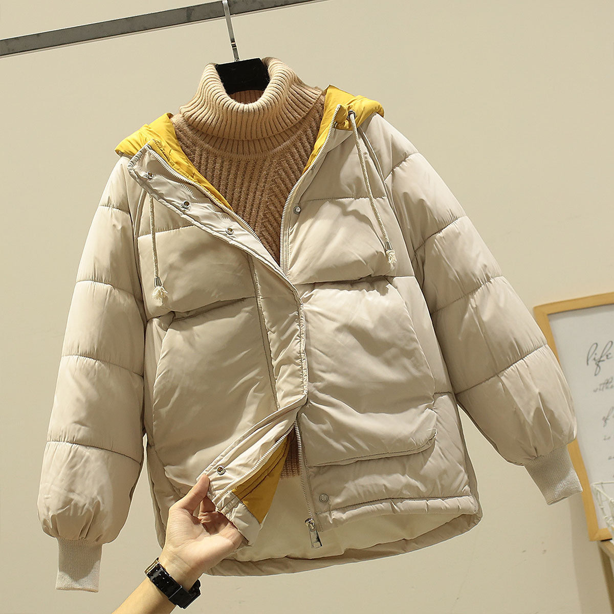 Giacca invernale imbottita beige con cappuccio giallo, chiusura a zip, tasche frontali, su sfondo neutro. Moda donna, abbigliamento caldo.