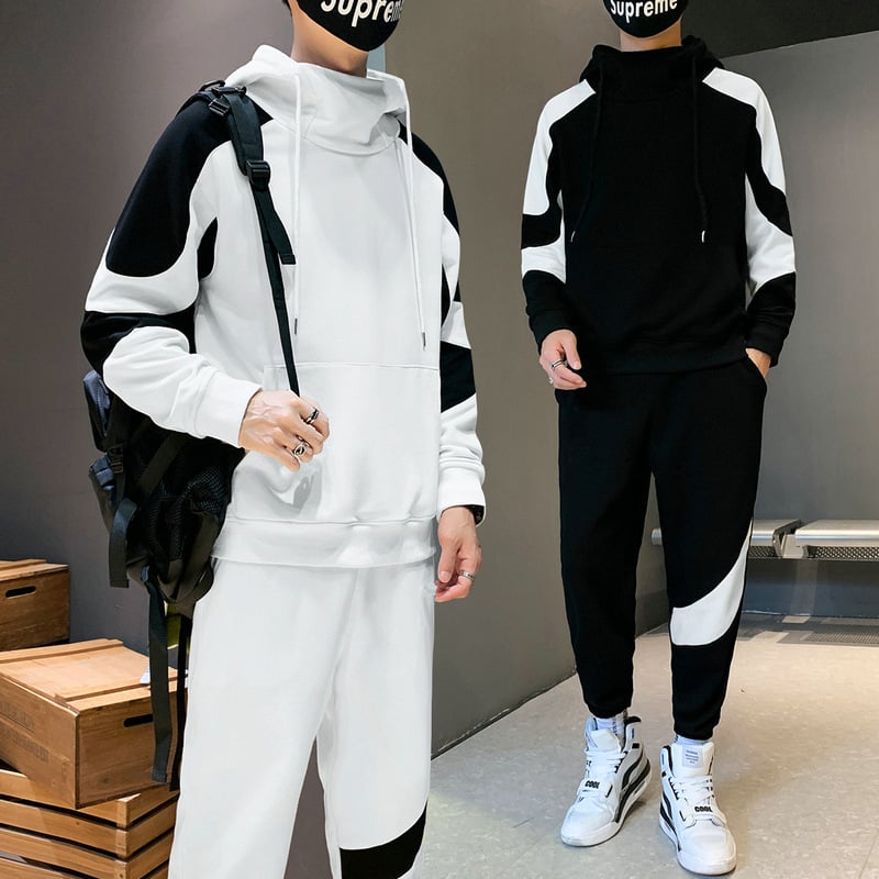 Due persone indossano tute sportive bianche e nere con cappuccio, scarpe da ginnastica bianche e maschere nere. Sfondo con scatole di legno.