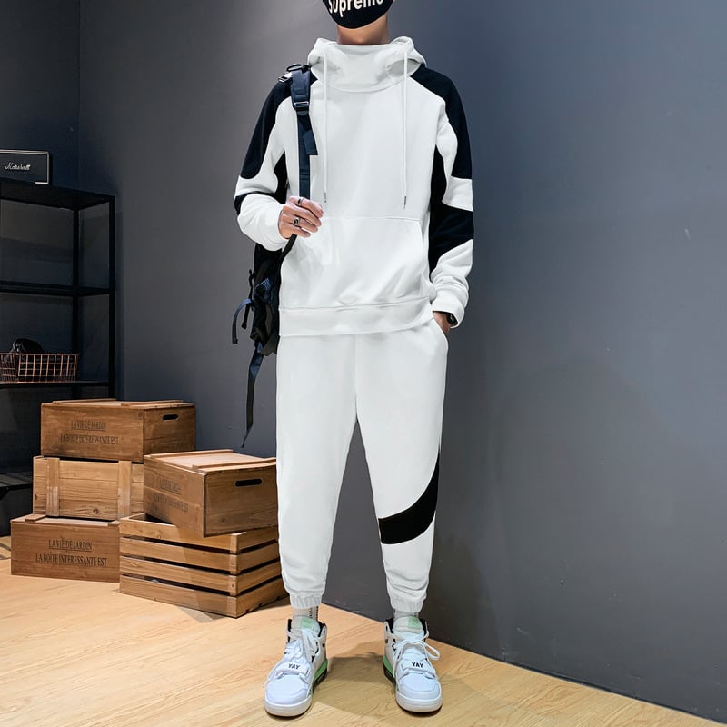 Uomo in tuta sportiva bianca e nera con cappuccio, zaino e scarpe da ginnastica, posa in una stanza moderna con scatole di legno.