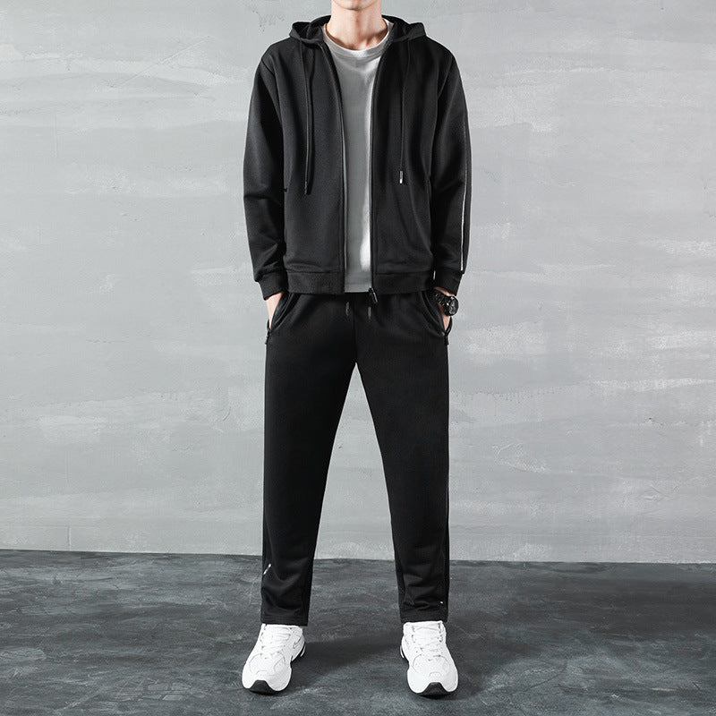 Uomo in tuta sportiva nera con felpa e pantaloni, indossa scarpe da ginnastica bianche, sfondo grigio. Abbigliamento casual, moda maschile.