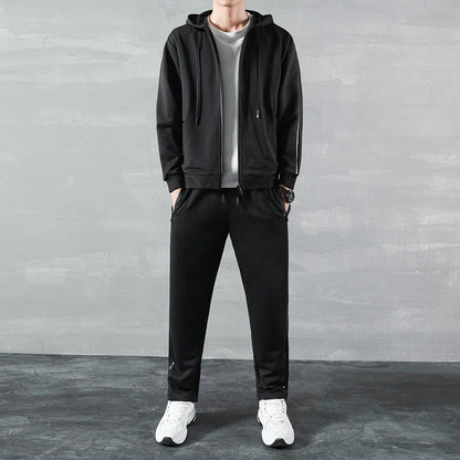 Uomo in tuta sportiva nera con felpa e pantaloni, indossa scarpe da ginnastica bianche, sfondo grigio. Abbigliamento casual, moda maschile.