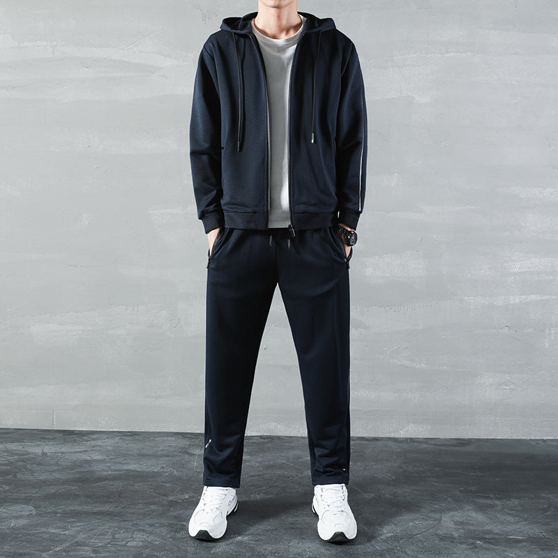 Uomo in tuta sportiva nera con felpa con cappuccio, t-shirt bianca e scarpe da ginnastica bianche, su sfondo grigio. Moda casual maschile.
