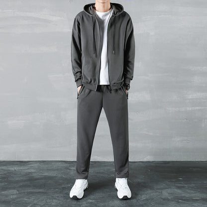 Uomo in tuta sportiva grigia con cappuccio, maglietta bianca e scarpe da ginnastica bianche, su sfondo grigio. Abbigliamento casual e comodo.