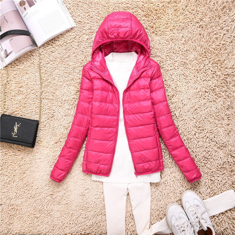 Giacca invernale rosa con cappuccio su tappeto beige, abbinata a maglia bianca e pantaloni chiari. Accessori: borsa nera e scarpe bianche.
