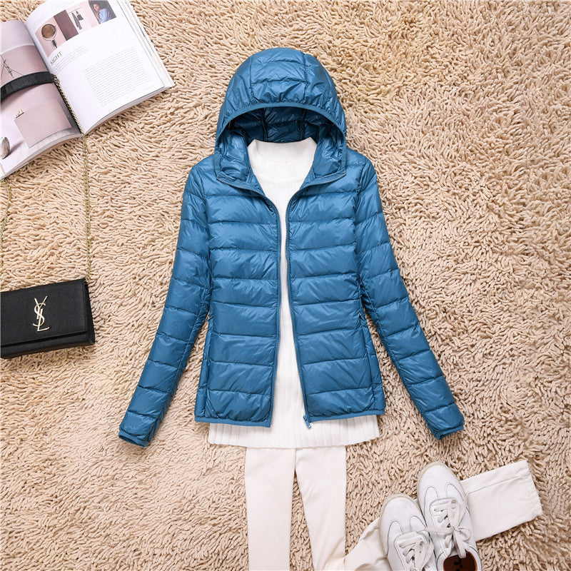 Giacca piumino blu con cappuccio su tappeto beige, abbinata a maglia bianca e pantaloni chiari. Accessori: borsa nera, scarpe bianche, rivista aperta.