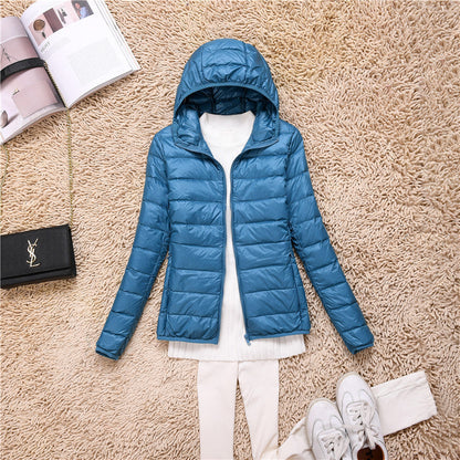 Giacca piumino blu con cappuccio su tappeto beige, abbinata a maglia bianca e pantaloni chiari. Accessori: borsa nera, scarpe bianche, rivista aperta.