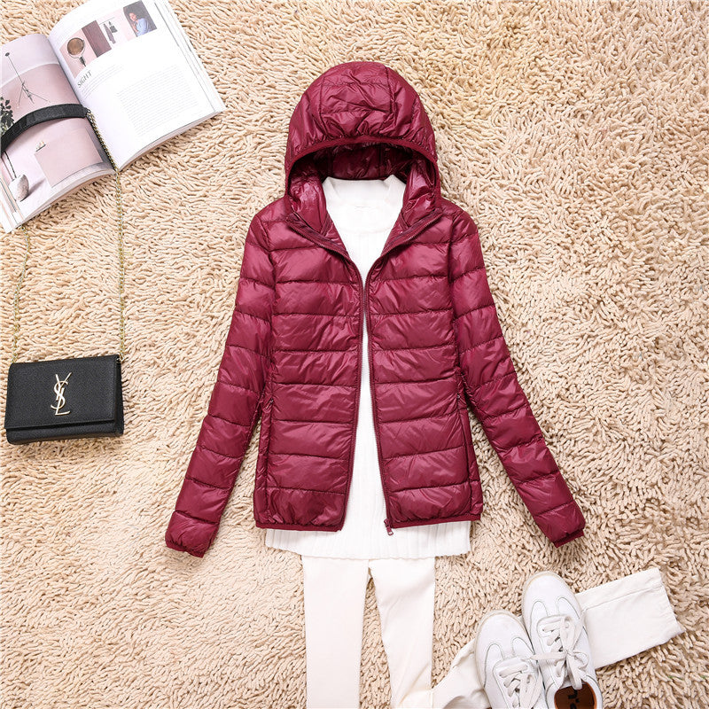Giacca piumino rosso bordeaux con cappuccio su tappeto beige, abbinata a maglia bianca, pantaloni chiari e scarpe da ginnastica, accanto a borsa nera e rivista.