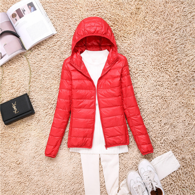 Giacca piumino rosso con cappuccio su tappeto beige, abbinata a maglia bianca e pantaloni chiari. Accessori: borsa nera e rivista aperta.