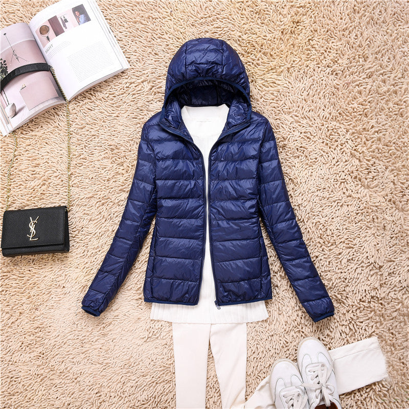 Giacca piumino blu con cappuccio su tappeto beige, abbinata a maglia bianca e scarpe da ginnastica. Accessori: borsa nera e rivista aperta.