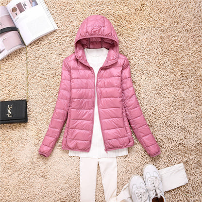 Giacca piumino rosa con cappuccio su tappeto beige, abbinata a maglia bianca e pantaloni chiari. Scarpe sportive bianche e borsa nera a lato.