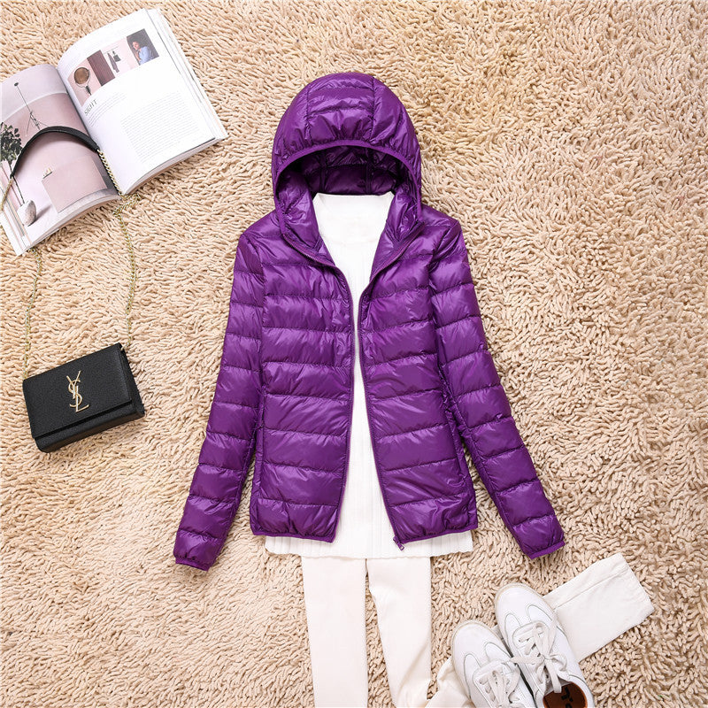 Giacca invernale viola con cappuccio su tappeto beige, abbinata a maglia bianca e pantaloni chiari. Accessori: borsa nera e rivista aperta.
