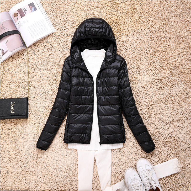 Giacca invernale nera con cappuccio su tappeto beige, abbinata a maglia bianca e scarpe sportive. Accessori: borsa nera e rivista aperta.