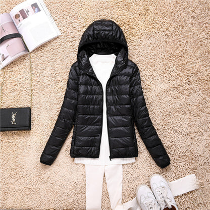 Giacca invernale nera con cappuccio su tappeto beige, abbinata a maglia bianca e scarpe sportive. Accessori: borsa nera e rivista aperta.