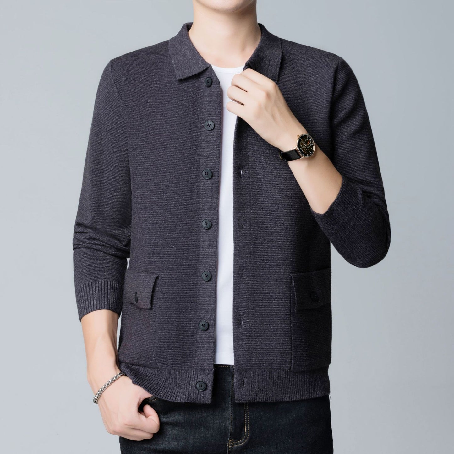 Uomo in cardigan grigio scuro con bottoni, indossa orologio nero e braccialetto argento, su sfondo grigio chiaro. Moda maschile elegante.