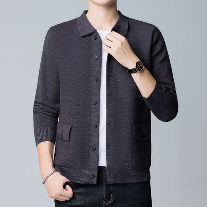 Uomo in cardigan grigio scuro con bottoni, indossa orologio nero e braccialetto argento, su sfondo grigio chiaro. Moda maschile elegante.