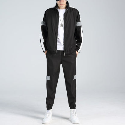 Uomo in tuta sportiva nera con strisce bianche, indossa scarpe da ginnastica bianche e collana, su sfondo grigio chiaro. Moda casual maschile.