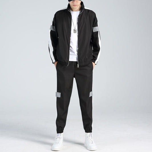 Uomo in tuta sportiva nera con strisce bianche, indossa scarpe da ginnastica bianche e collana, su sfondo grigio chiaro. Moda casual maschile.