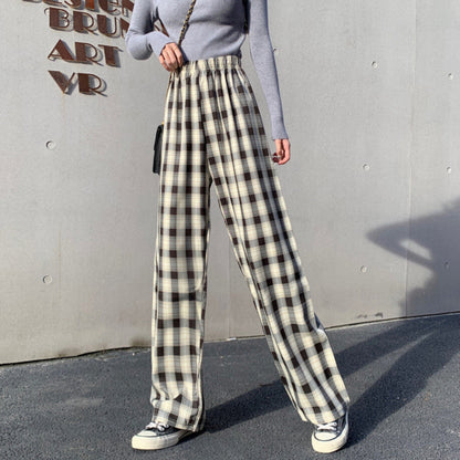 Donna con pantaloni a quadri bianchi e neri, maglia grigia a maniche lunghe, borsa a tracolla nera, scarpe da ginnastica bianche. Sfondo grigio.