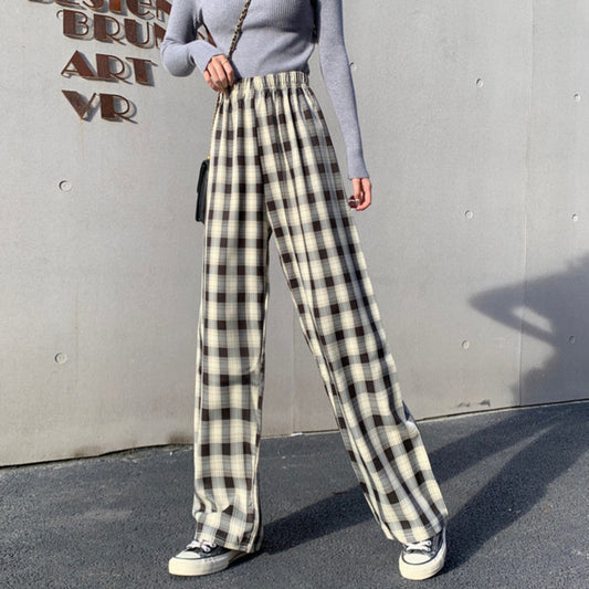 Donna con pantaloni a quadri bianchi e neri, maglia grigia a maniche lunghe, borsa a tracolla nera, scarpe da ginnastica bianche. Sfondo grigio.