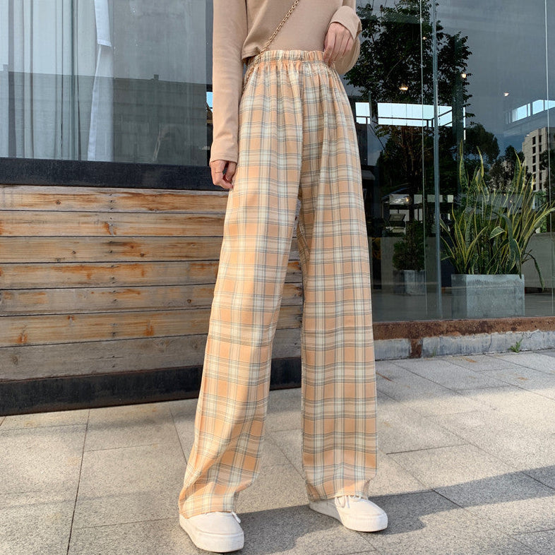 Pantaloni a quadri beige e arancioni, stile casual, indossati con maglia beige e scarpe bianche, su sfondo urbano con vetrina e piante.