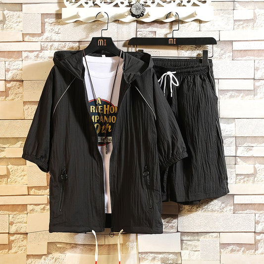 Completo sportivo nero da uomo con giacca a vento e pantaloncini, appeso su grucce. Sfondo in mattoni chiari. Abbigliamento casual estivo.