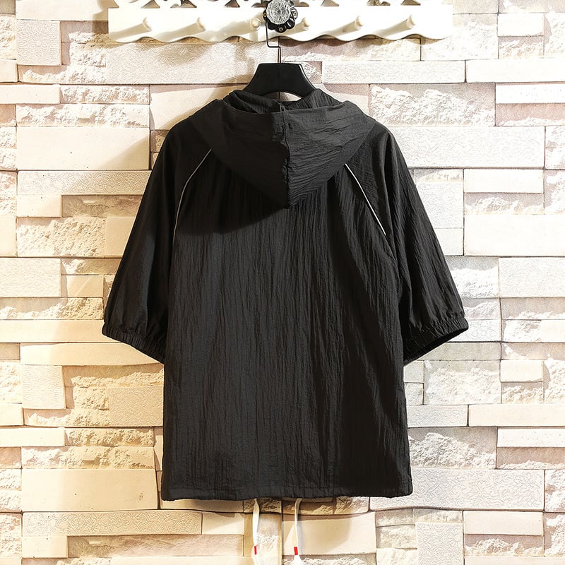 Giacca a vento nera con cappuccio, maniche corte, appesa a un appendiabiti su sfondo di mattoni chiari. Design casual, ideale per la moda urbana.