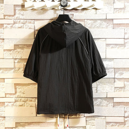 Giacca a vento nera con cappuccio, maniche corte, appesa a un appendiabiti su sfondo di mattoni chiari. Design casual, ideale per la moda urbana.