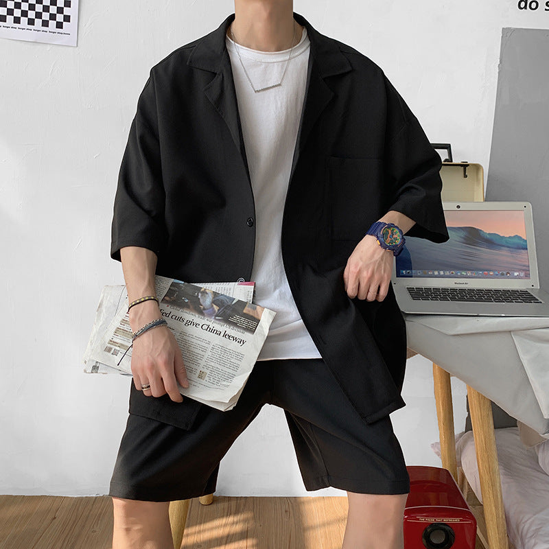 Uomo in abbigliamento casual nero con camicia bianca, tiene un giornale. Laptop su tavolo in sfondo. Moda estiva, stile urbano, outfit moderno.