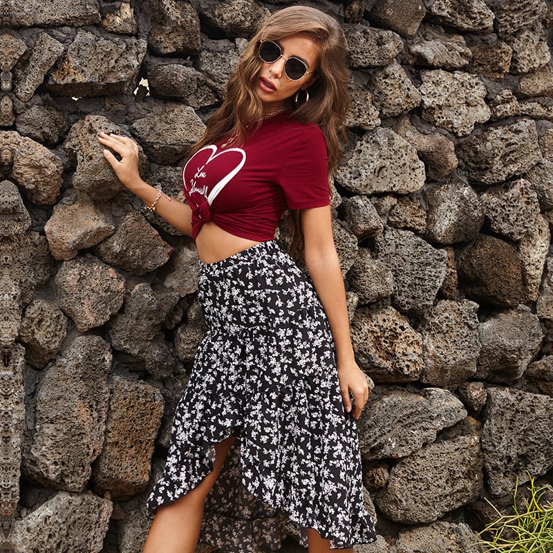 Donna con occhiali da sole indossa maglietta rossa con cuore e gonna asimmetrica floreale bianca e nera, posa davanti a muro di pietra. Moda estiva casual.