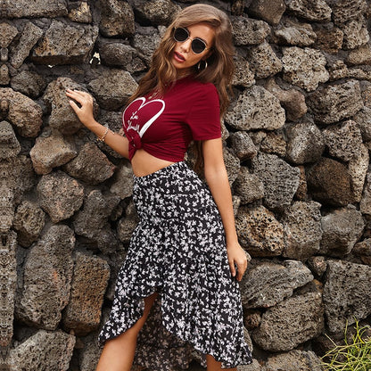 Donna con occhiali da sole indossa maglietta rossa con cuore e gonna asimmetrica floreale bianca e nera, posa davanti a muro di pietra. Moda estiva casual.