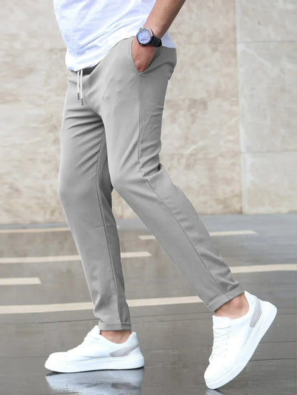 Uomo con pantaloni grigi eleganti e scarpe da ginnastica bianche, cammina su marciapiede bagnato. Orologio nero al polso, stile casual moderno.