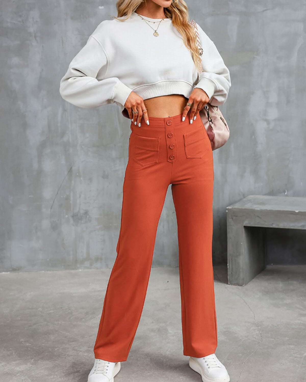 Donna con maglione bianco corto e pantaloni a vita alta arancioni, posa in piedi su sfondo grigio. Moda casual, abbigliamento autunnale.