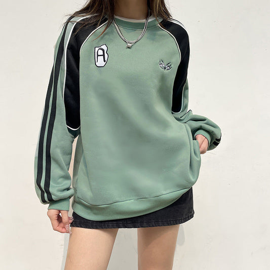 Felpa verde con dettagli neri e strisce sulle maniche, indossata con gonna nera. Design casual, perfetta per moda streetwear e look autunnali.