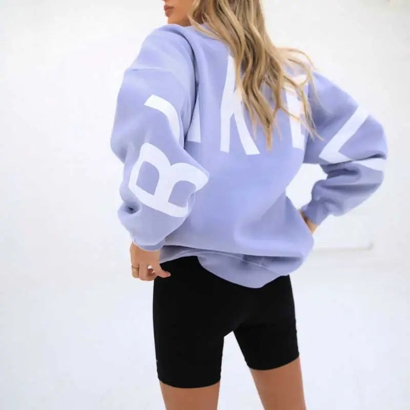 Donna con felpa oversize lilla con scritta bianca, pantaloncini neri. Moda casual, abbigliamento sportivo donna, tendenze streetwear.